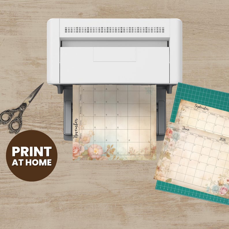 a printer, a pair of scissors, and two calendar pages with floral designs, all placed on a wooden surface.