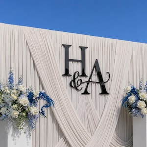 May include: A large event backdrop featuring cream-coloured pleated fabric draped in a crisscross pattern. Centered on the fabric are large, glossy black letters H and A with an ampersand. Two floral arrangements with blue and white flowers sit below.