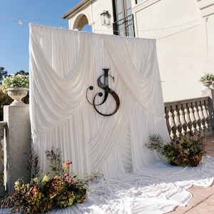 May include: A white fabric backdrop featuring draped curtains and a large, black, stylised letter JS monogram mounted in the centre. The fabric pools on the ground, and the display is accented by floral arrangements with green and brown tones.
