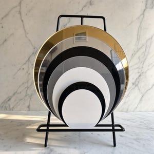May include: A set of nested circular acrylic discs in varying sizes and colours, including gold, black, white, and translucent grey, arranged in a stack. The discs are held upright by a black metal wire display stand with a rectangular base.