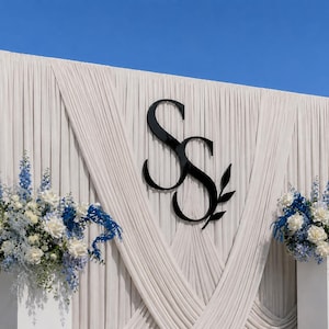 May include: A large event backdrop featuring draped off-white fabric with a prominent black, glossy, interlocking SS monogram logo centred. Two white pedestals flank the display, each topped with floral arrangements of blue and white flowers.