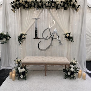 May include: A wedding backdrop featuring a white tufted bench, a white rug, and white sheer curtains. A large silver mirrored monogram with the letters L and A is centred. The display is decorated with white and dark green floral arrangements.