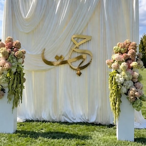 May include: A white fabric backdrop with draped folds features a large gold-coloured Arabic calligraphy sign in the centre. Two white rectangular pedestals stand on either side, each topped with a large arrangement of pink, white, and green flowers.