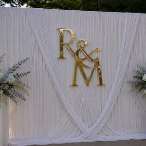 May include: A large white fabric backdrop featuring draped curtains and a central gold metallic monogram sign displaying the letters R and M connected by an ampersand. Two white pedestals flank the display, each topped with white floral arrangements.