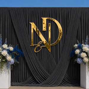 May include: A dark grey fabric backdrop featuring a large, reflective gold monogram of the letters N and D with an ampersand. Two white rectangular pedestals stand on either side, each topped with a floral arrangement of blue and white flowers.