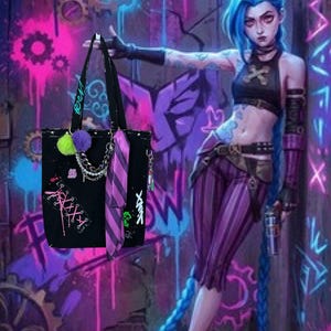May include: A black tote bag featuring a pink lace-up graphic, a purple and green pom-pom charm, a silver chain detail, and a purple and black striped ribbon. The bag has black straps and is displayed against a vibrant graffiti-covered wall background.