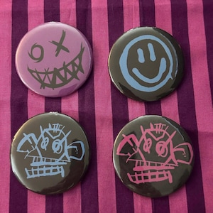 May include: Four circular pinback buttons. The top left is purple with a dark gray grinning face. The top right is black with a light blue smiley face. The bottom left is black with a light blue abstract character. The bottom right is black with a pink abstract character.