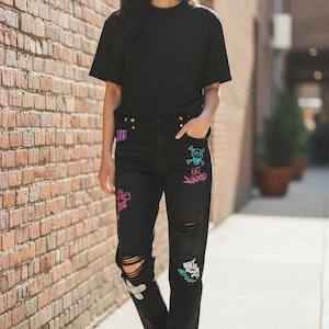 Puede incluir: Camiseta negra y vaqueros negros desgastados con dise&ntilde;os coloridos de estilo grafiti. Los vaqueros tienen rodillas rasgadas y se combinan con zapatillas blancas. El atuendo lo lleva una persona caminando por una acera.