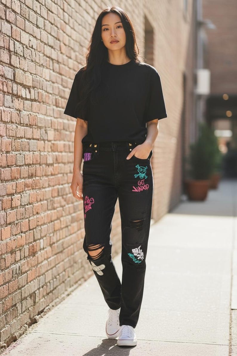 Puede incluir: Camiseta negra y vaqueros negros desgastados con dise&ntilde;os coloridos de estilo grafiti. Los vaqueros tienen rodillas rasgadas y se combinan con zapatillas blancas. El atuendo lo lleva una persona caminando por una acera.