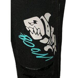 Puede incluir: Tela negra con un dise&ntilde;o de esqueleto de tibur&oacute;n blanco y la palabra "BOOM" en turquesa. El tibur&oacute;n tiene dientes afilados y una expresi&oacute;n amenazante. La tela tiene un aspecto desgastado.