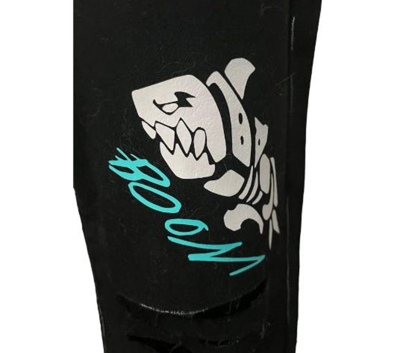 Puede incluir: Tela negra con un dise&ntilde;o de esqueleto de tibur&oacute;n blanco y la palabra "BOOM" en turquesa. El tibur&oacute;n tiene dientes afilados y una expresi&oacute;n amenazante. La tela tiene un aspecto desgastado.