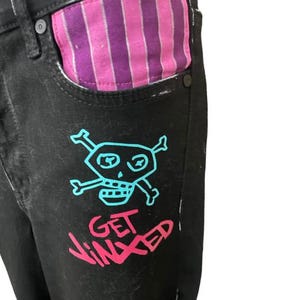 Puede incluir: Vaqueros de mezclilla negros con un gr&aacute;fico de calavera y huesos cruzados turquesa y el texto "GET JINXED" en rosa. El forro del bolsillo presenta un patr&oacute;n de rayas rosas y moradas. Una declaraci&oacute;n de moda &uacute;nica y atrevida.