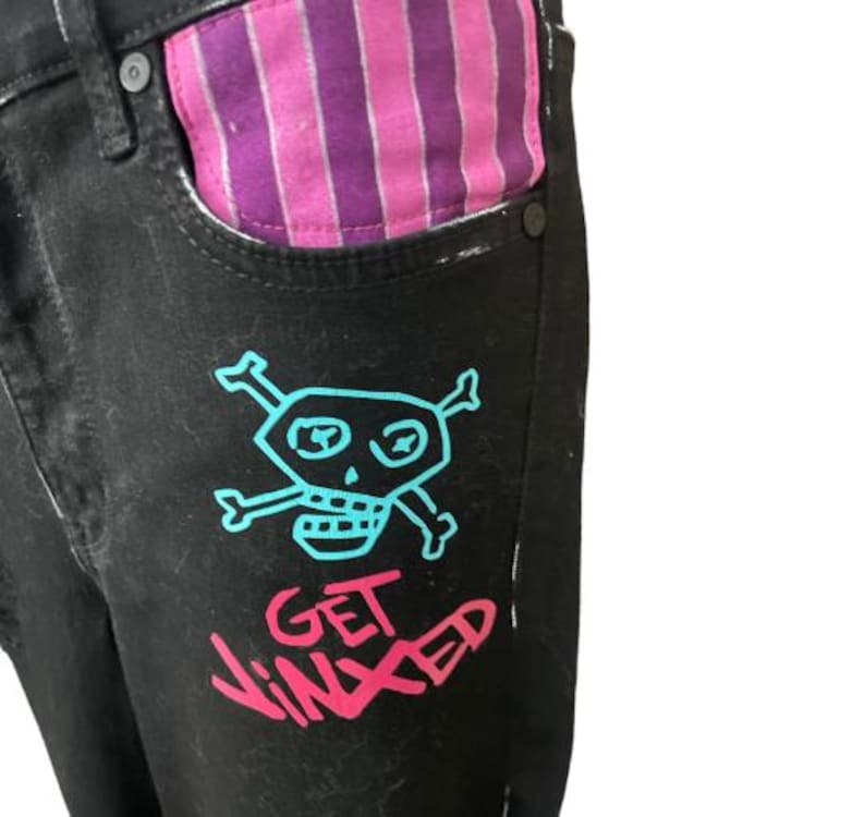 Puede incluir: Vaqueros de mezclilla negros con un gr&aacute;fico de calavera y huesos cruzados turquesa y el texto "GET JINXED" en rosa. El forro del bolsillo presenta un patr&oacute;n de rayas rosas y moradas. Una declaraci&oacute;n de moda &uacute;nica y atrevida.