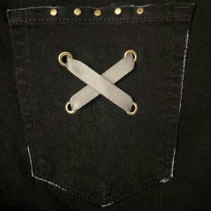 Puede incluir: Primer plano de un bolsillo de mezclilla negro con un dise&ntilde;o decorativo. El bolsillo presenta un detalle cruzado de cinta gris asegurado con ojales dorados. El borde superior est&aacute; adornado con peque&ntilde;os tachuelas doradas.