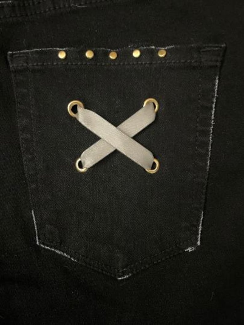 Puede incluir: Primer plano de un bolsillo de mezclilla negro con un dise&ntilde;o decorativo. El bolsillo presenta un detalle cruzado de cinta gris asegurado con ojales dorados. El borde superior est&aacute; adornado con peque&ntilde;os tachuelas doradas.