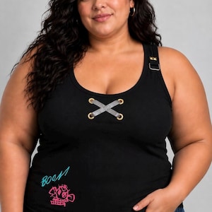 May include: A black sleeveless tank top featuring a scoop neckline with gold-toned grommets and a grey ribbon lace-up detail. The left shoulder strap has a gold-toned buckle, and the lower front displays the word BOOM in blue and a pink graphic.