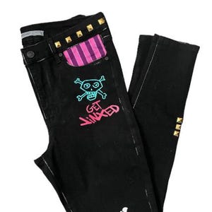 Puede incluir: Vaqueros negros con un gr&aacute;fico de calavera y huesos cruzados y el texto "GET JINXED" en rosa. Los vaqueros tienen un bolsillo a rayas rosa y morado y tachuelas doradas.