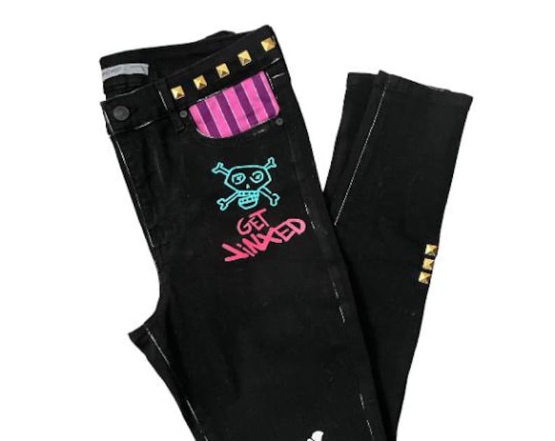 Puede incluir: Vaqueros negros con un gr&aacute;fico de calavera y huesos cruzados y el texto "GET JINXED" en rosa. Los vaqueros tienen un bolsillo a rayas rosa y morado y tachuelas doradas.