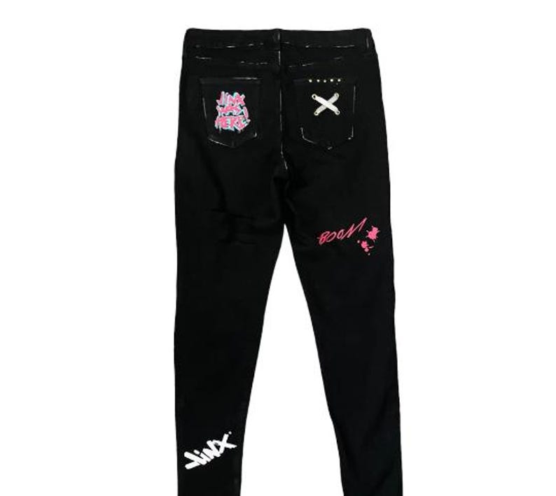 Puede incluir: Pantalones vaqueros negros con textos y dise&ntilde;os de estilo graffiti en blanco y rosa. Los bolsillos traseros presentan un dise&ntilde;o de "X" y un gr&aacute;fico rosa y blanco. La palabra "Jinx" est&aacute; escrita en blanco en la parte inferior izquierda de la pierna.