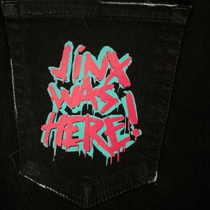Puede incluir: Bolsillo de mezclilla negro con el texto "JINX WAS HERE!" en un dise&ntilde;o estilo grafiti. El texto es de color rosa brillante con un contorno turquesa, creando un contraste audaz contra la tela oscura.