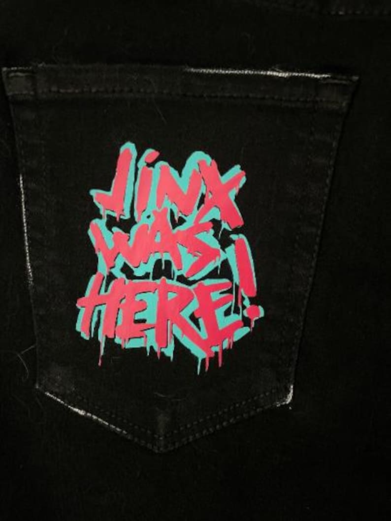 Puede incluir: Bolsillo de mezclilla negro con el texto "JINX WAS HERE!" en un dise&ntilde;o estilo grafiti. El texto es de color rosa brillante con un contorno turquesa, creando un contraste audaz contra la tela oscura.