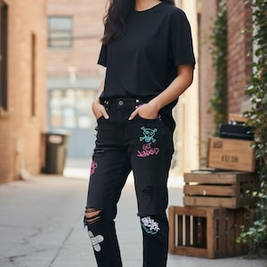 Puede incluir: Camiseta negra y vaqueros negros desgastados con parches coloridos de calaveras y flores. Los vaqueros tienen rodillas rasgadas y se combinan con zapatillas blancas. El texto "GET JINXED" es visible en los vaqueros.