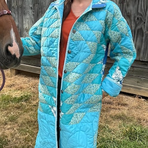 May include: A long, quilted coat in shades of turquoise and blue, featuring a geometric pattern and floral accents. The coat has a button-up front and a collar. The sleeves are long, and the coat reaches the ankles. The coat is worn outdoors.