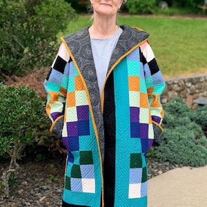 May include: A long-sleeved quilted coat featuring a patchwork design of square fabric blocks in shades of turquoise, purple, orange, green, and light blue. The coat has a dark gray patterned hood and trim, with a mustard yellow edge binding.