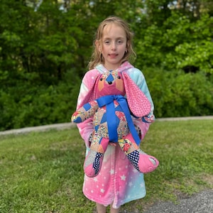 May include: A handmade patchwork bunny rabbit with pink, blue, and floral patterned fabric. The bunny has long pink ears and a blue ribbon around its neck. The bunny is being held by a person wearing a pink, blue, and white tie-dye robe.