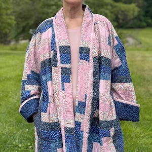 May include: A patchwork quilted jacket in shades of pink and blue. The jacket has a shawl collar and pockets. The sleeves are cuffed with a solid blue fabric. The jacket is worn by a person. The design incorporates floral patterns and geometric shapes.