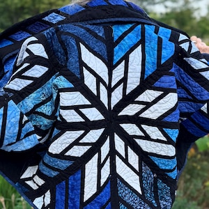 May include: A quilted jacket featuring a large, central star pattern in shades of white, light blue, and deep navy blue. The design is constructed from various fabric textures and patterns, outlined with black fabric strips and intricate stitching.