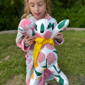 May include: A handmade white bunny plush toy with green, red, and yellow accents. The bunny has a yellow ribbon around its neck and is being held by a person wearing a colorful robe. The bunny's ears and feet have a red floral pattern.