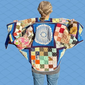 May include: A patchwork quilt jacket with a blue border and a large white doily design in the center. The jacket features a variety of colorful fabric squares and patterns. The person is wearing blue jeans.