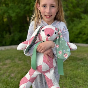 May include: A handmade stuffed bunny with a patchwork design in pink and white. The bunny has long ears with a floral pattern and a green ribbon. The bunny is being held by a person.