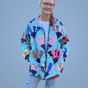 May include: A quilted jacket featuring a fan-shaped patchwork design in shades of blue, pink, and various patterned fabrics. The jacket has a button-up front and a collar. The person is wearing a white t-shirt and blue jeans.