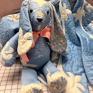 May include: A stuffed rabbit toy crafted from blue and cream quilted fabric with floral patterned accents on the ears and feet. The rabbit features black button eyes, a black nose, and a solid pink ribbon tied around its neck, positioned next to a matching blue and cream quilted blanket.