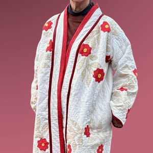 May include: A white quilted coat with red trim and floral appliqués. The coat features a wrap-around design with a red border along the edges and down the front. Red flowers are appliquéd across the coat.