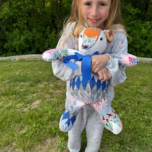 May include: A handmade patchwork teddy bear with a blue ribbon. The bear features a variety of colorful fabric patterns, including floral, geometric, and solid colors. The bear's head is white with orange and brown accents. The bear is being held by a person.