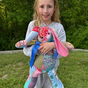 May include: A handmade patchwork bunny rabbit doll with a blue ribbon. The doll features a variety of colorful fabric patterns, including floral, paisley, and plaid. The bunny has long, floppy ears and is being held by a person.