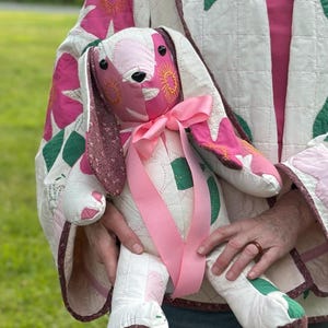 May include: A handmade stuffed bunny with pink and white patchwork design, a pink ribbon, and embroidered details. The bunny is held in front of a quilted jacket with floral patterns.