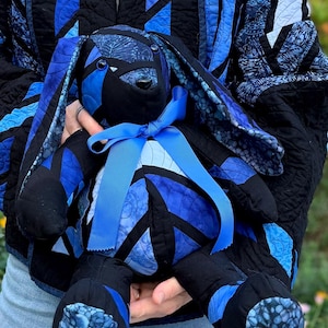 May include: A stuffed rabbit toy constructed from patchwork fabric in various shades of blue and black. The rabbit features long, floppy ears, a rounded body, and a bright blue satin ribbon tied in a bow around its neck. The fabric patterns include solid black, mottled blue, and swirling leaf designs.