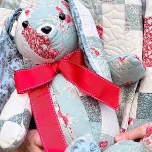 May include: A stuffed rabbit toy crafted from quilted fabric featuring a patchwork of light blue, white, and red floral patterns. The rabbit has black button eyes, a black stitched nose, and a bright red ribbon tied in a bow around its neck.