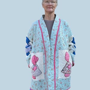 May include: A light blue quilted jacket with a floral pattern and pink trim. The jacket features two pockets with appliqued figures and patchwork details on the sleeves. The jacket is approximately 102 cm long.