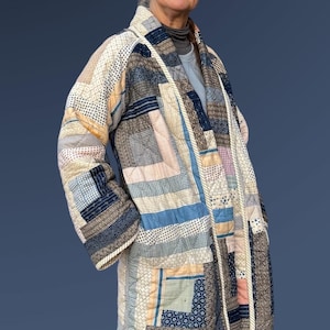 May include: A patchwork quilted coat in shades of blue, beige, and white. The coat features a variety of geometric patterns and textures, with a rope trim along the edges. The coat is knee-length and has long sleeves.