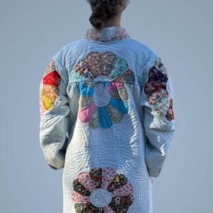 May include: A light blue quilted coat with floral patchwork details. The coat features large, colorful flower appliqués on the back and sleeves, with a quilted pattern throughout. The collar and cuffs have a floral print.