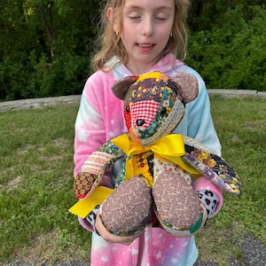 May include: A handmade patchwork teddy bear with a yellow ribbon around its neck. The bear is made from various fabric patterns, including floral, gingham, and solid colors. The bear's ears and paws are a solid brown color.