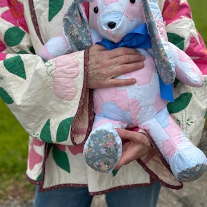 May include: A handmade patchwork bunny rabbit in pastel pink, blue, and white. The bunny has long ears with floral fabric accents and a blue ribbon. The bunny is being held by someone wearing a floral quilted jacket.