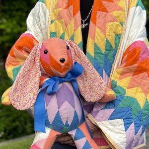 May include: A colorful patchwork bunny plush toy with a blue ribbon, held in front of a person wearing a vibrant, geometric-patterned quilt jacket. The bunny features pink, orange, and purple hues.