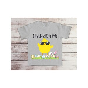 May include: A gray t-shirt with a yellow chick wearing sunglasses and the text "Chicks Dig Me". The chick is standing on a bed of green grass with colorful Easter eggs.
