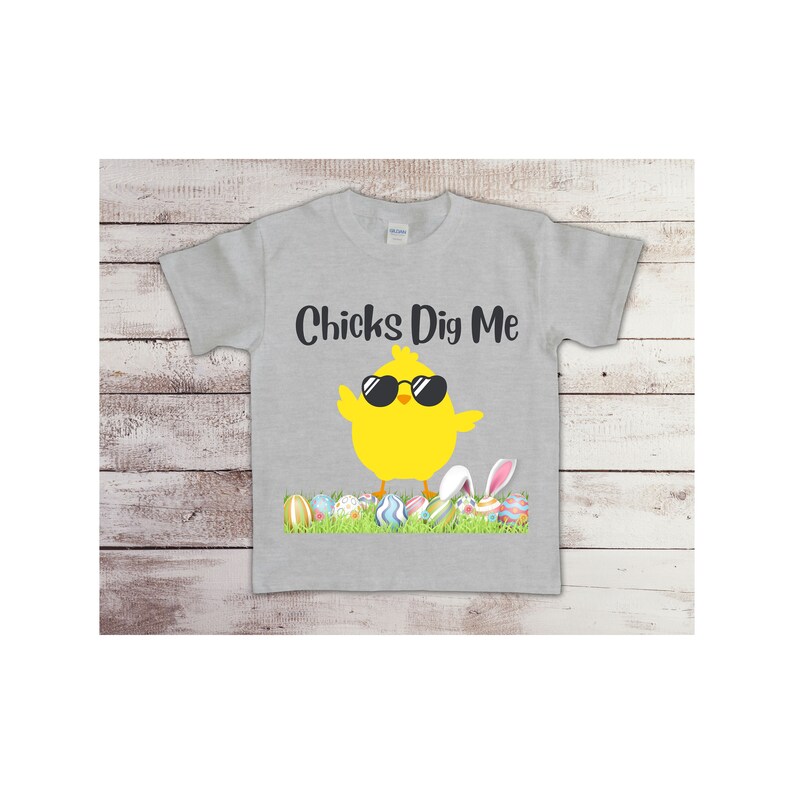 May include: A gray t-shirt with a yellow chick wearing sunglasses and the text "Chicks Dig Me". The chick is standing on a bed of green grass with colorful Easter eggs.